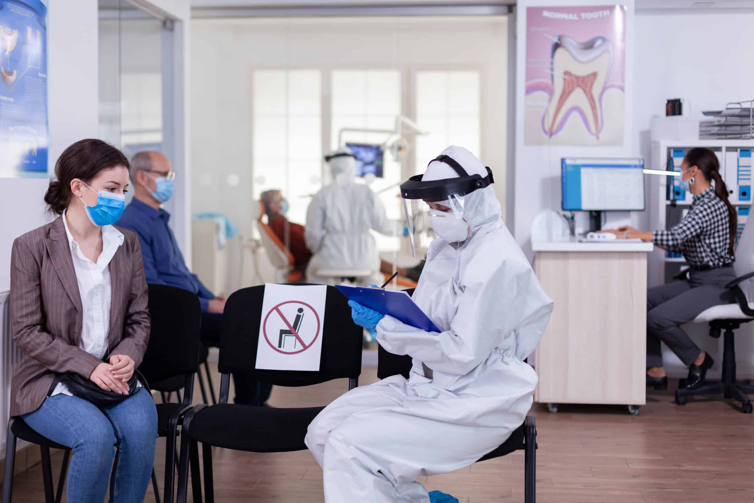 Dentist assistant with ppe equipment talking with patient before consultation during coronavirus epidemic sitting on chairs in waiting area keeping distance